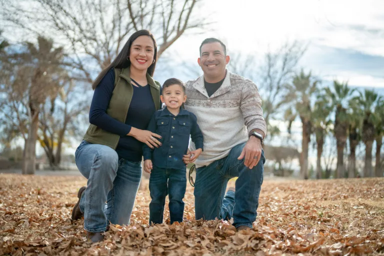 Familia de tres arrodillada junta al aire libre en un parque, sonriendo a la cámara
