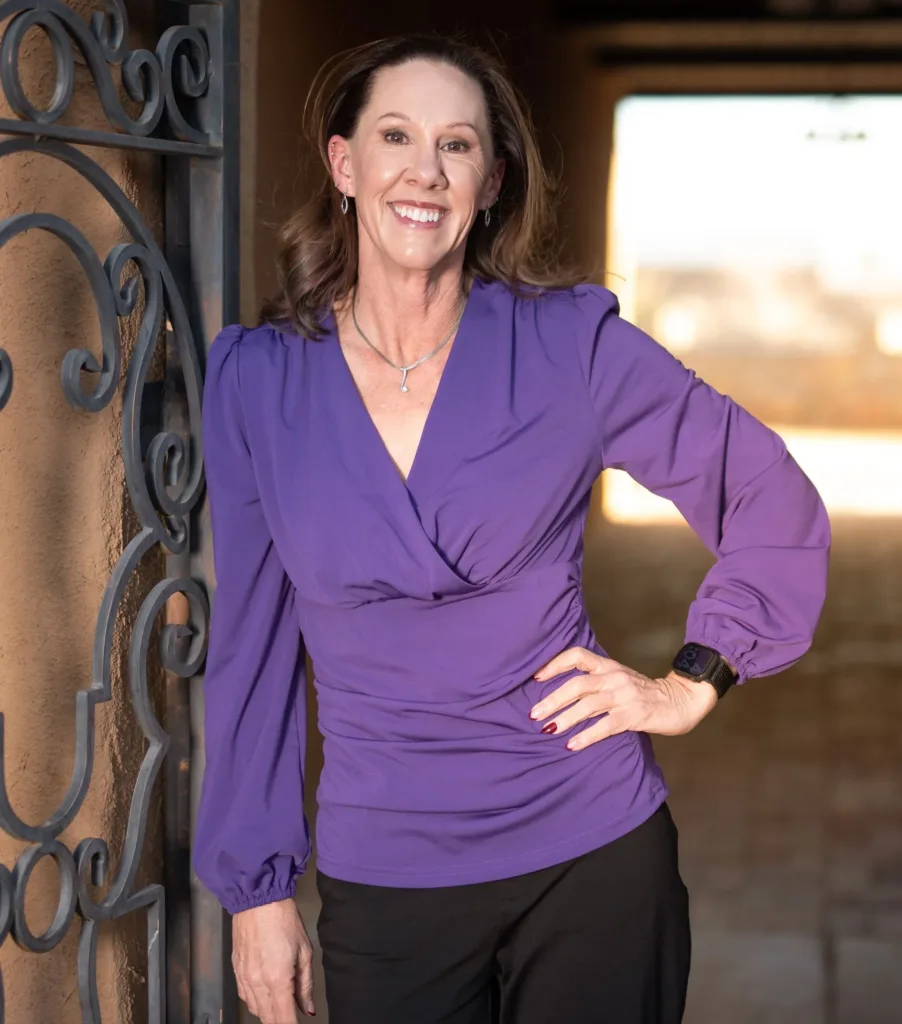 A smiling woman in a purple long-sleeved shirt stands against a stucco wall with a decorative metal gate.
