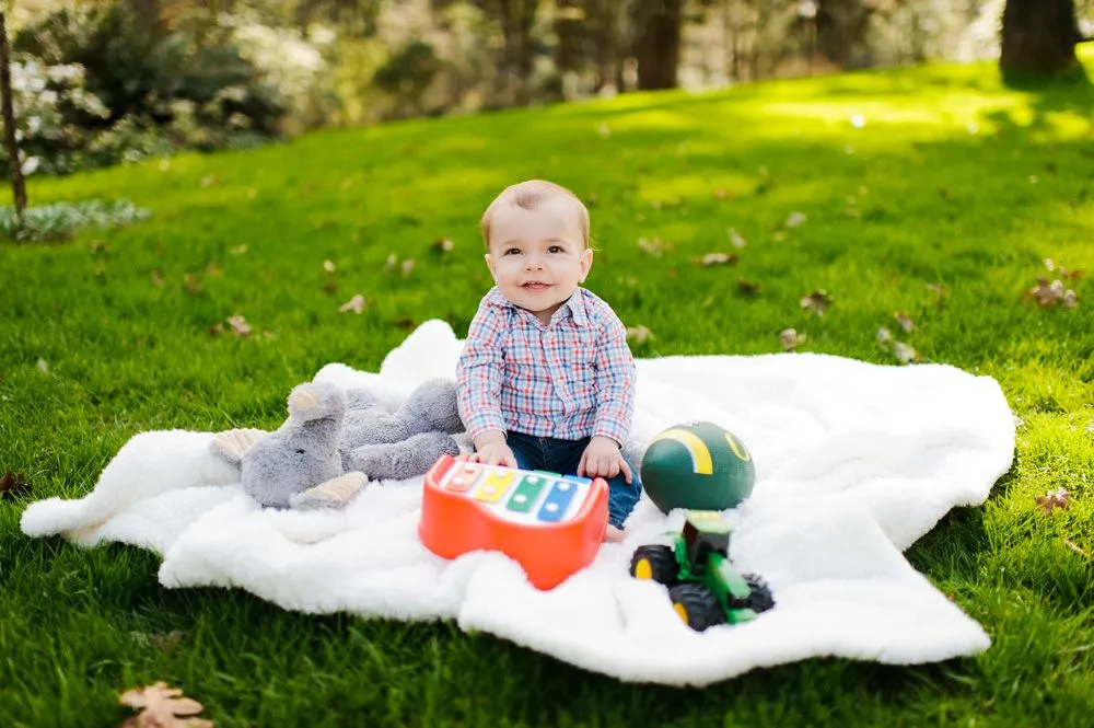 Baby sitting on a white blanket in a park, smiling at the camera, with toys.