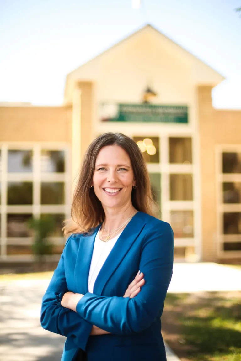 Woman in blue blazer, arms crossed, smiles in front of a tan building.