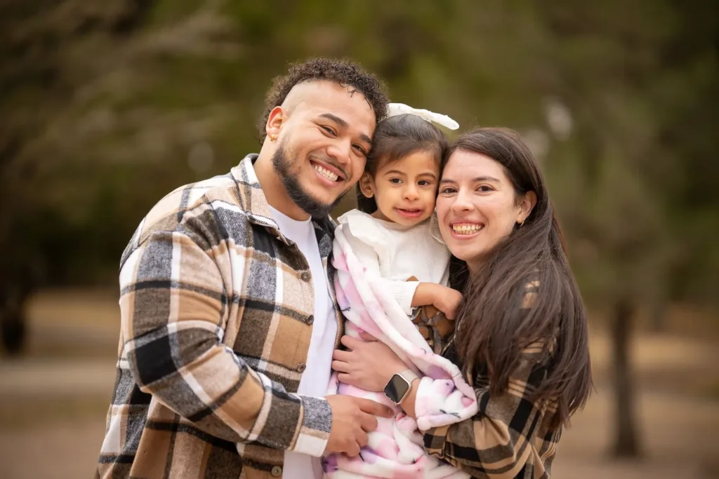 A smiling family holding their young child in an outdoor setting.