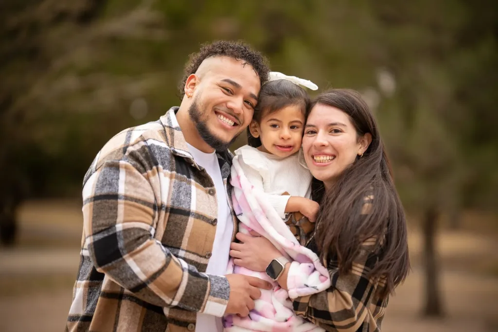 Smiling family of three outdoors, holding a baby in a wooded park.