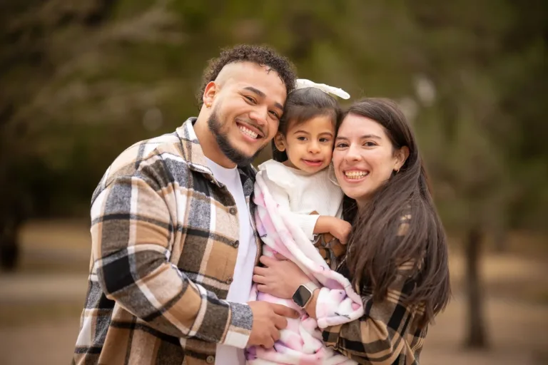 Smiling family of three outdoors, holding a baby in a wooded park.