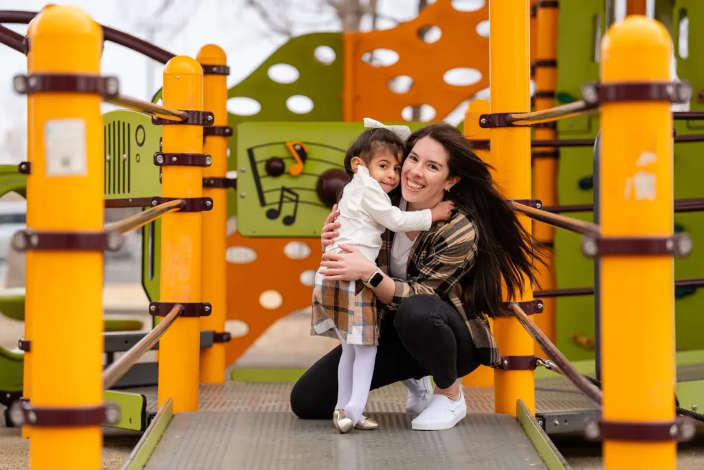 Mujer abrazando a un niño en un colorido parque infantil. Están sonriendo. El parque tiene estructuras naranjas y verdes.