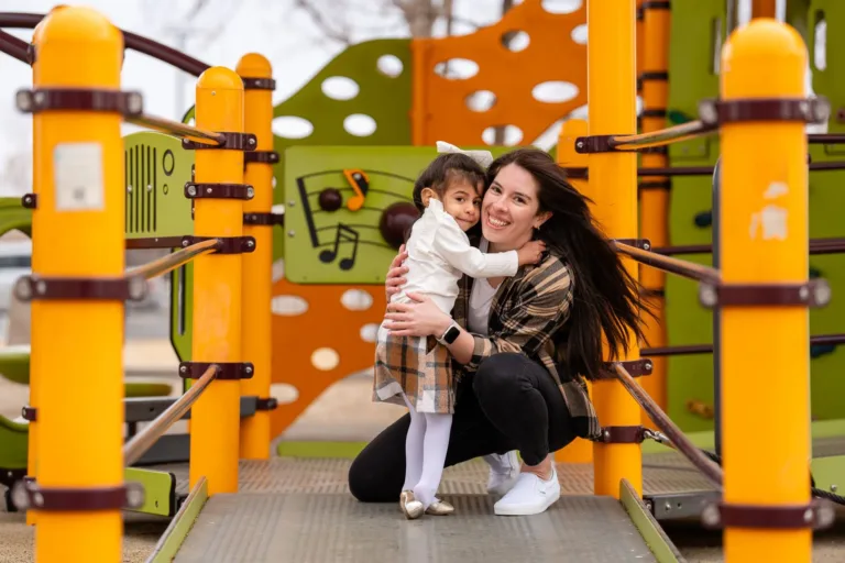 Mujer abrazando a un niño en un colorido parque infantil. Están sonriendo. El parque tiene estructuras naranjas y verdes.