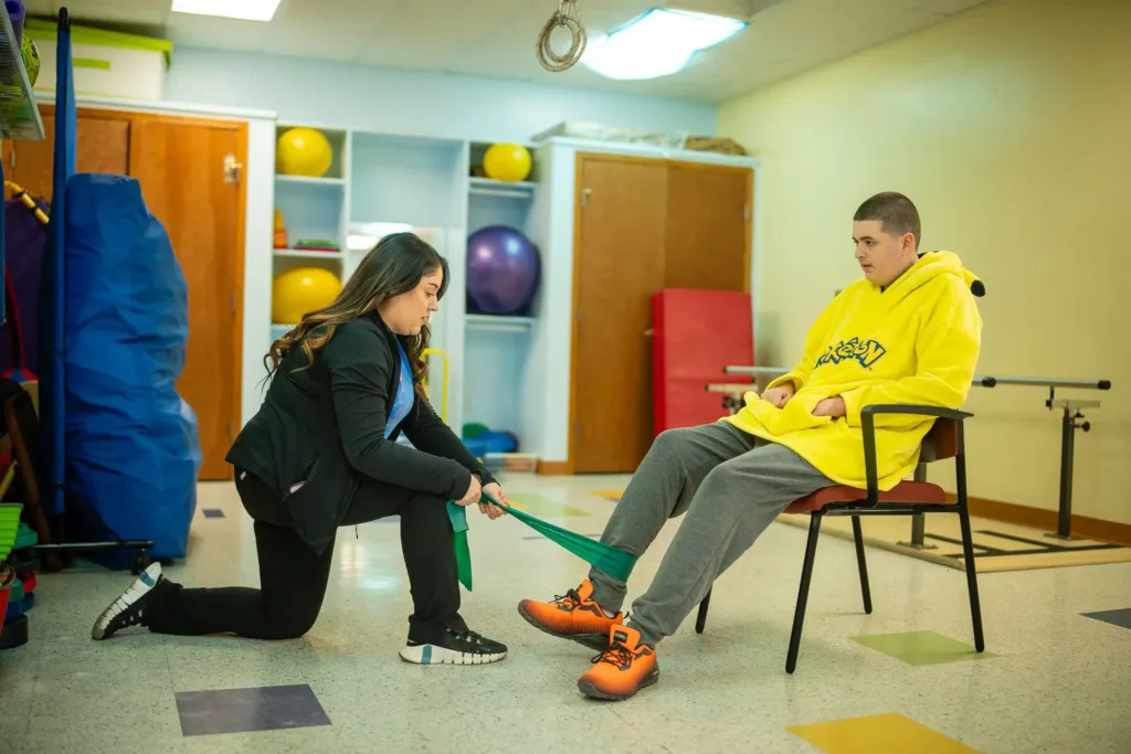 Fisioterapeuta pediátrico arrodillado junto a un niño sentado, asistiendo con ejercicios de piernas.
