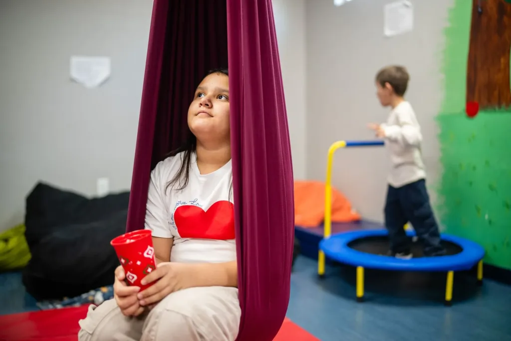 Girl sits in purple swing, holding a red cup, looking upwards. Boy bounces on a small trampoline in the background.