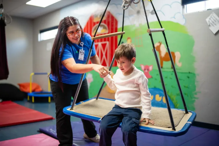 Terapeuta asiste a un niño sentado en un columpio. El niño está en una sala de terapia con un mural.