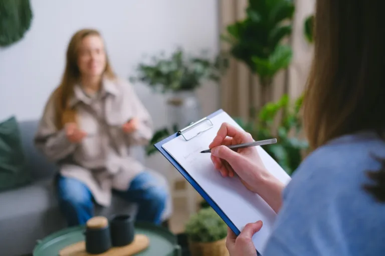 Person taking notes on clipboard while speaking with another person on a couch.