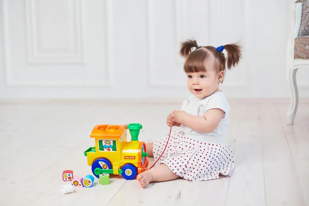 Girl with pigtails sits and plays with a yellow toy train on a white floor.