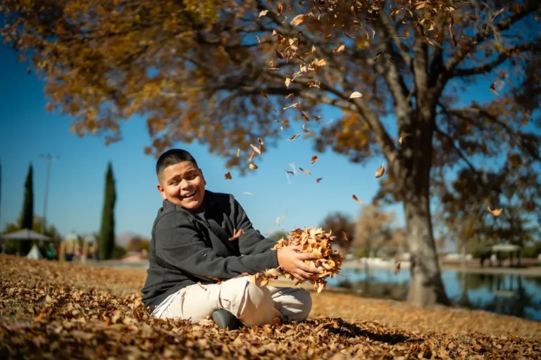 Niño sentado en hojas de otoño las lanza al aire cerca de un lago.