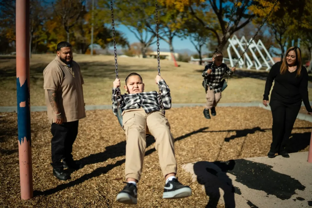 Family at a playground: child on swing, parents watching, another child swings in the background. Sunny day.