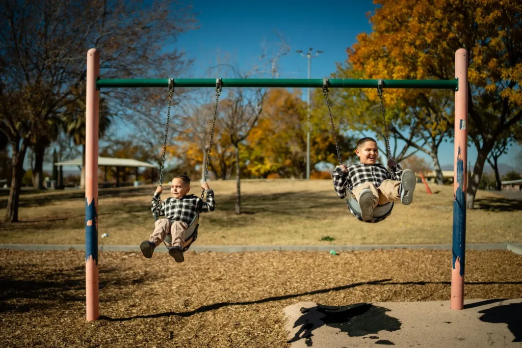 Two children swinging on swings in a park with fall foliage in the background.