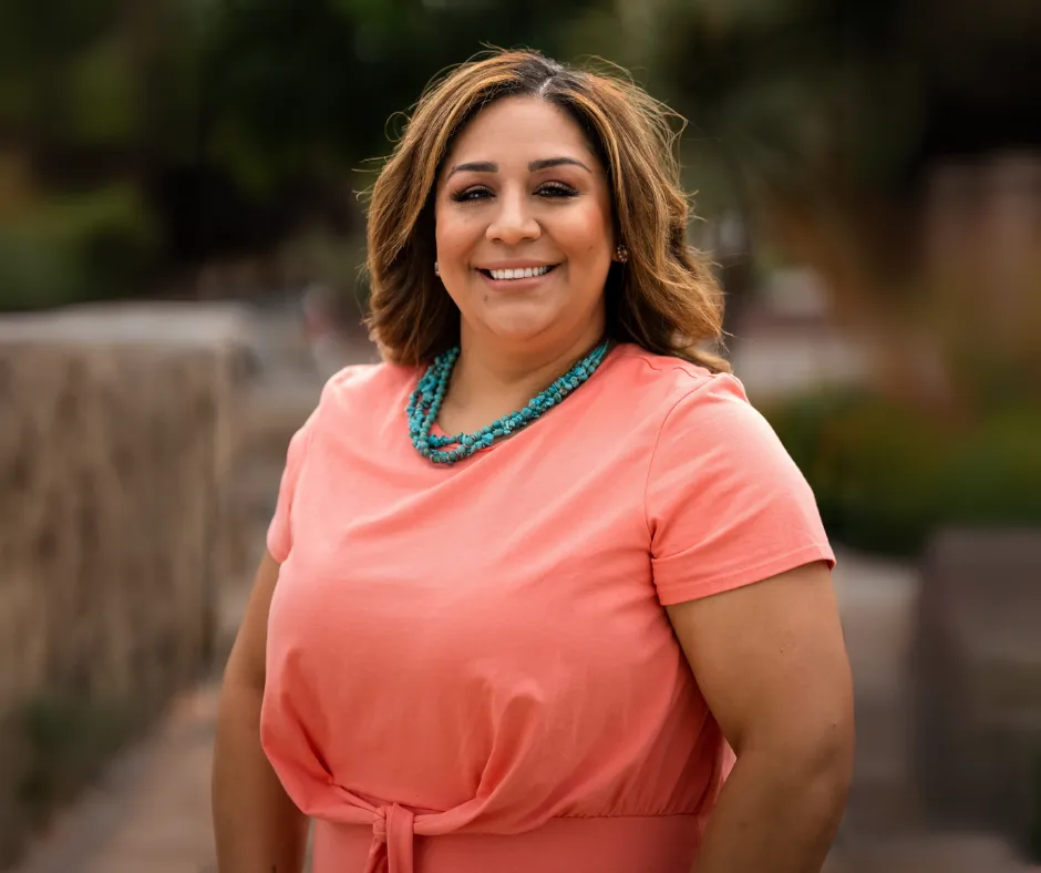 Woman wearing a coral dress and turquoise necklace, smiling outdoors.
