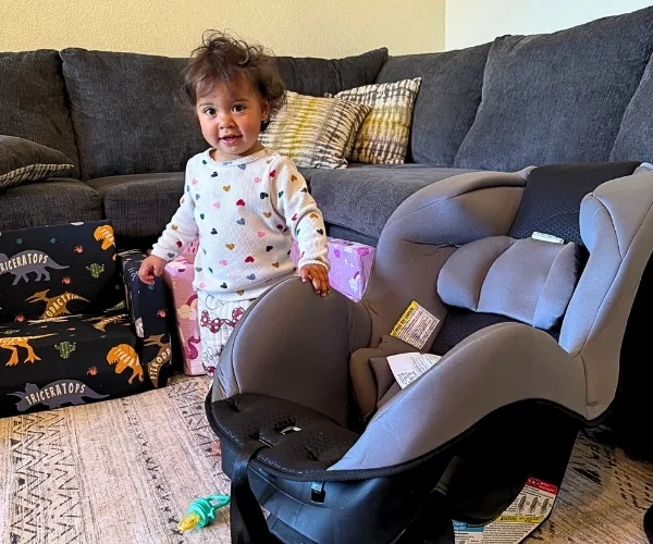 Niño pequeño de pie junto a una silla de auto gris en una sala de estar