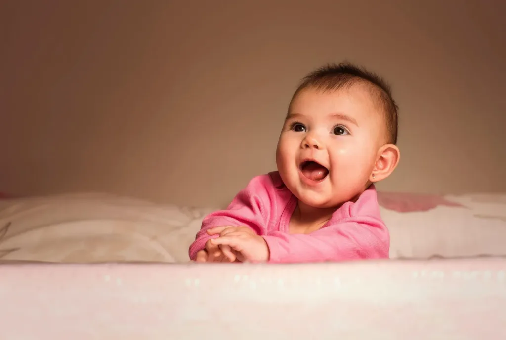 Baby in pink outfit, laughing, leaning on a white bed with pink floral accents, warm lighting.
