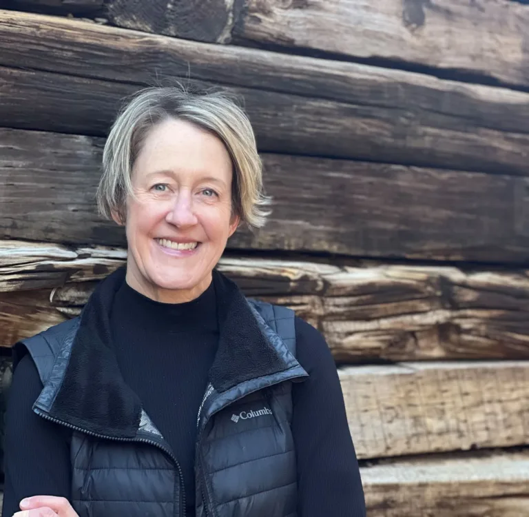 Dr. Williams with blonde hair smiles, wearing a black vest and sweater, set against a wooden wall.