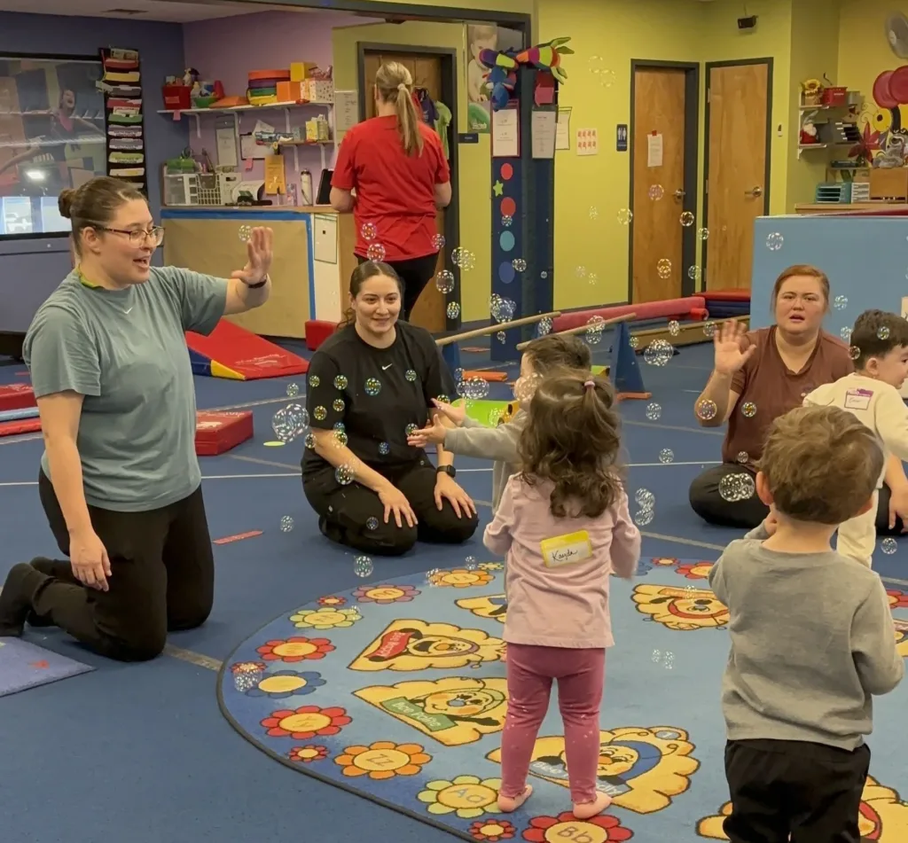 Three adults and three children interact with bubbles in a brightly colored indoor play area with a patterned rug.
