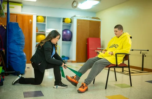 Terapeuta ayuda a un joven sentado a ejercitar su pierna con una banda de resistencia en una sala de rehabilitación