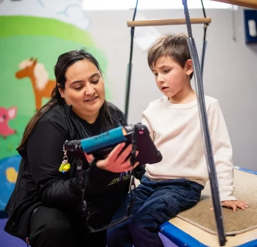 Terapeuta mostrándole una tableta a un niño pequeño en un columpio de terapia