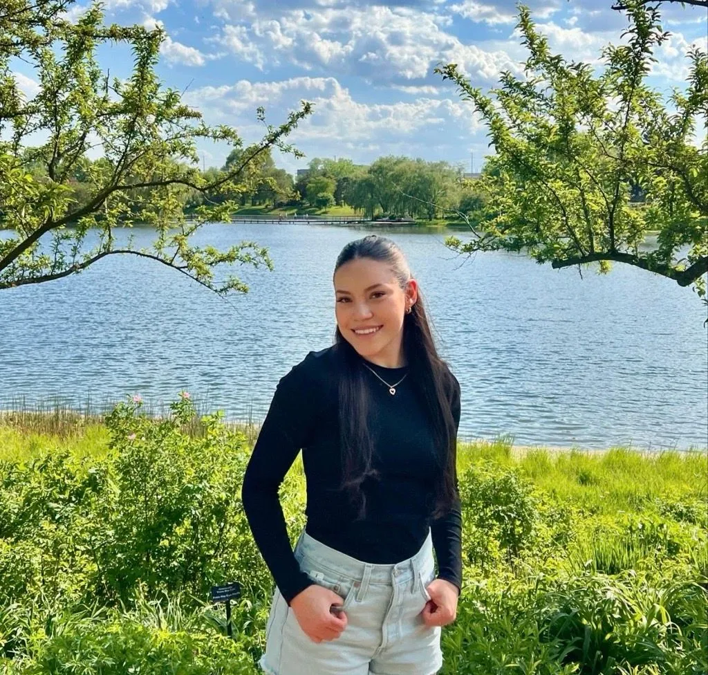 Kenia Portillo smiles outdoors in a black top and light shorts, standing by a lake with trees.