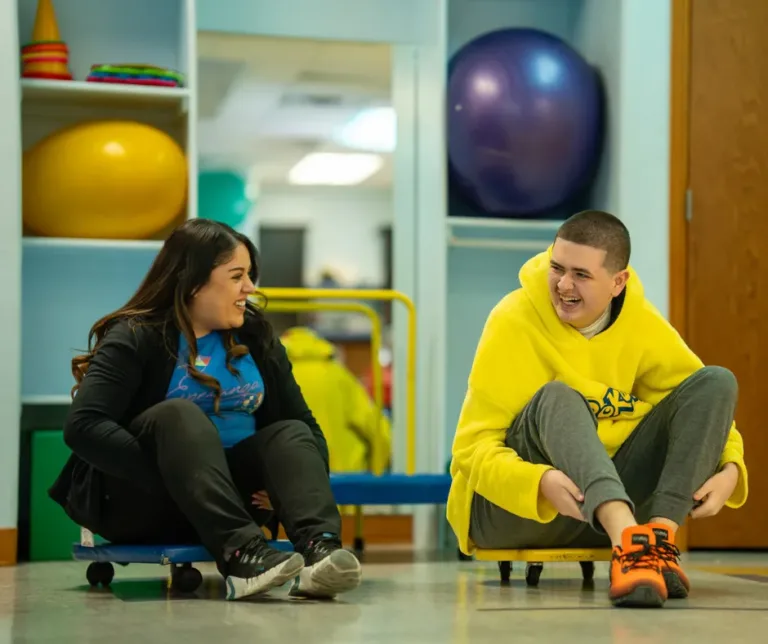 Woman and person in yellow hoodie sitting on wheeled boards, smiling at each other in a room with exercise equipment.