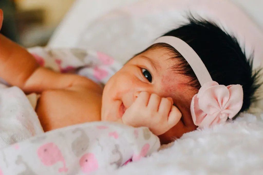 Newborn baby with dark hair and pink bow headband smiles. Lying on white blanket, hand near face.