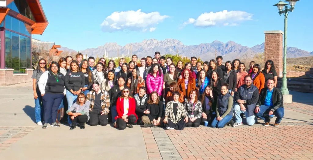 Large group of people posing outdoors; mountains in the background, sunny day.