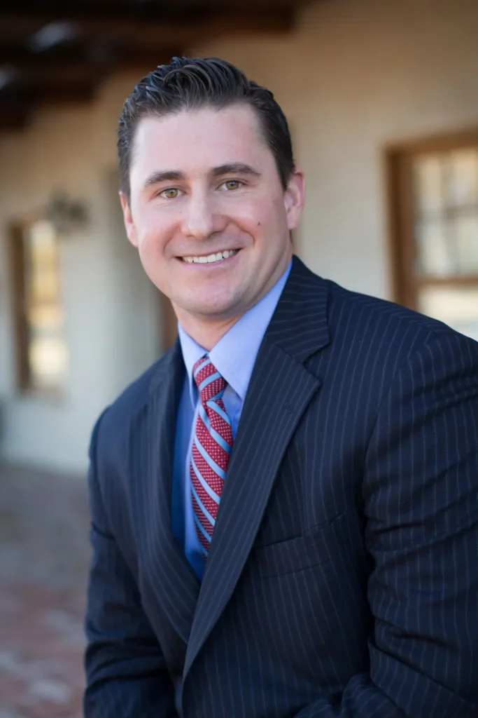 Man in suit smiling outdoors, leaning slightly forward.