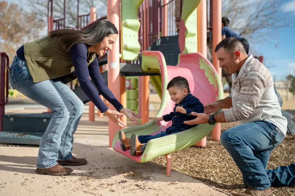 Smiling young boy on a swing, outdoors; blue shirt, jeans, brown shoes, sunny day. Mariposa Autism