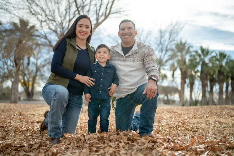 Family of three, smiling, kneeling in fallen leaves; trees in background.