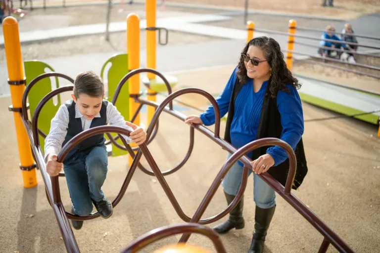 Una persona con una camisa azul ayuda a un niño en un obstáculo de un parque infantil.