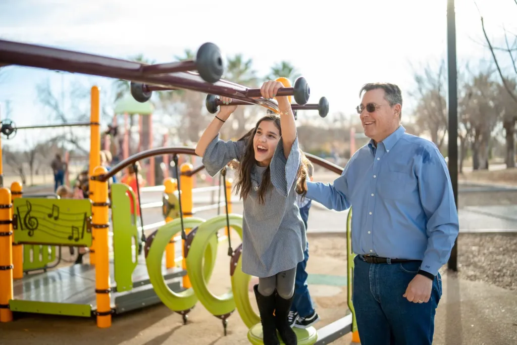 Girl swinging on monkey bars at a playground, with her dad smiling beside her, sunny day.