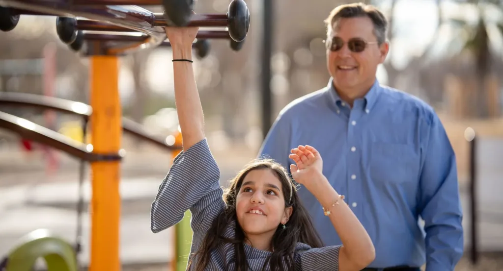 Girl on playground monkey bars, reaching up, smiling; man watching in background.
