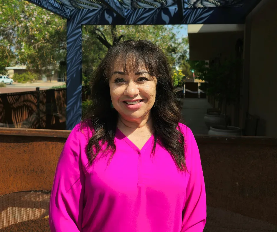 Woman in a pink shirt smiling outdoors, standing near a building with a blue trellis.