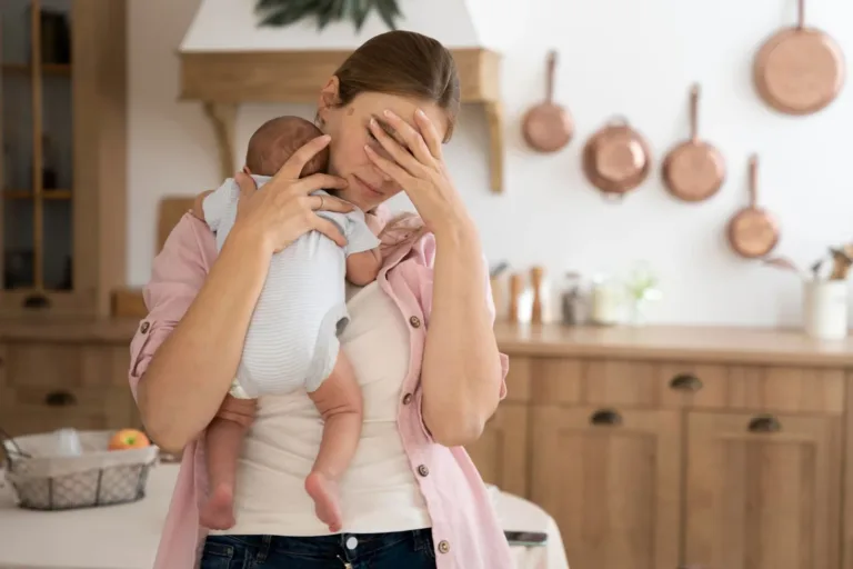Woman holding infant, looks stressed, hand on face, in a kitchen with copper pots.