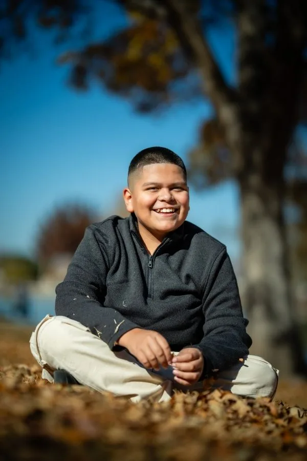 A smiling boy sitting cross-legged outdoors on fallen leaves